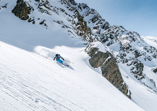 Ein Skifahrer in blauer Ausrüstung gleitet einen steilen, verschneiten Berghang mit felsigen Gipfeln im Hintergrund unter einem klaren Himmel hinunter.