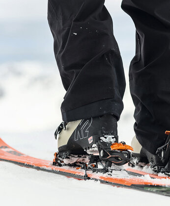 Nahaufnahme der Skischuhe einer Person, die in orangefarbene Skier auf verschneitem Boden eingeklinkt sind, wobei die Winterkleidung sichtbar ist.