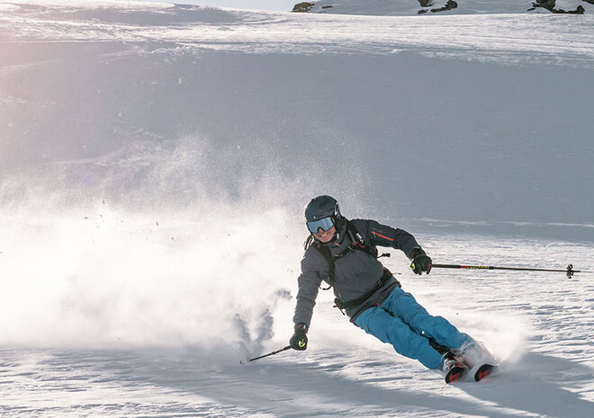 A snowboarder in blue pants glides down a snowy slope, with sunlit mountains in the background.