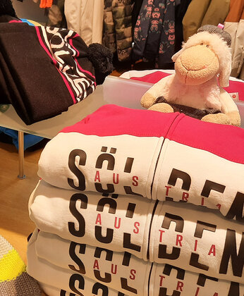 Vier gefaltete Sweatshirts mit der Aufschrift Sölden auf einem Tisch, auf dem ein lächelndes Plüschschaf sitzt.