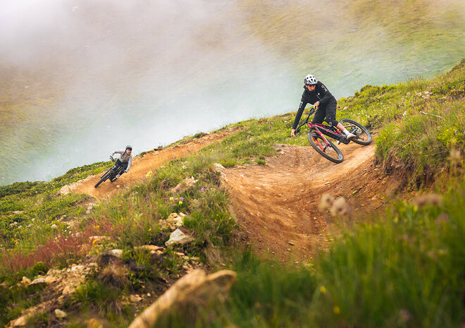 Two mountain bikers ride down a grassy mountainside under cloudy skies.