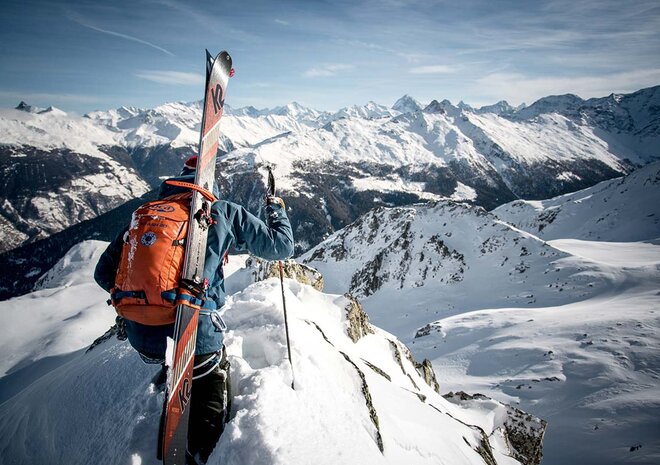 A ski tourer with a red backpack stands on a snow-covered ridge and looks out over a range of snow-covered peaks.