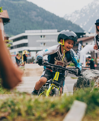 Mountain bikers riding on a practice course, with mountains in the background and spectators nearby.