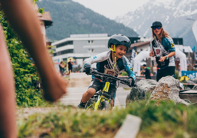 Mountainbiker, die auf einem Übungsgelänfe fahren, mit den Bergen im Hintergrund und Zuschauern in der Nähe.