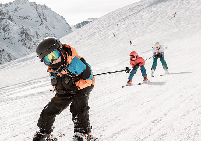 Three people are skiing down a snowy mountainside, the sky is clear, and snow-capped peaks can be seen in the distance.
