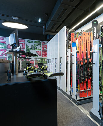 Innenraum eines Skiverleihs mit Ski- und Snowboardständern, Schuhregalen und farbenfroher Wandkunst im Hintergrund.