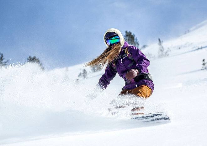 Person, die an einem klaren, sonnigen Tag mit einer lila Jacke, einem Helm und einer Skibrille auf einer verschneiten Piste Snowboard fährt.