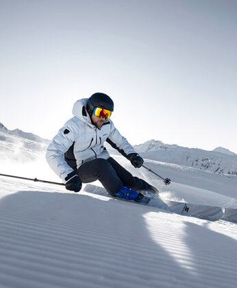 Ein Skifahrer in Winterausrüstung fährt an einem sonnigen Tag in einer verschneiten Berglandschaft durch frisch präparierten Schnee.