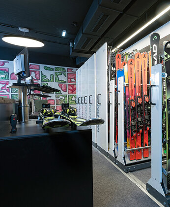 Innenraum eines Skiverleihs mit Ski- und Snowboardständern, Schuhregalen und farbenfroher Wandkunst im Hintergrund.