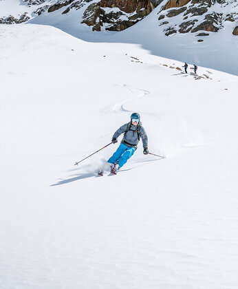 Ein Skifahrer in Blau fährt eine verschneite Piste mit Felsen und schneebedeckten Bergen im Hintergrund hinunter.