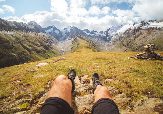 Person, die mit ausgestreckten Beinen auf einer grasbewachsenen Hügelkuppe sitzt, mit Blick auf die Berge und den wolkenverhangenen Himmel in der Ferne.