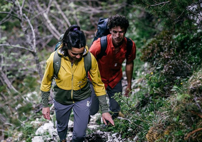 Zwei Personen mit Rucksäcken wandern einen felsigen, bewaldeten Pfad hinauf, der von Grün und Bäumen umgeben ist.