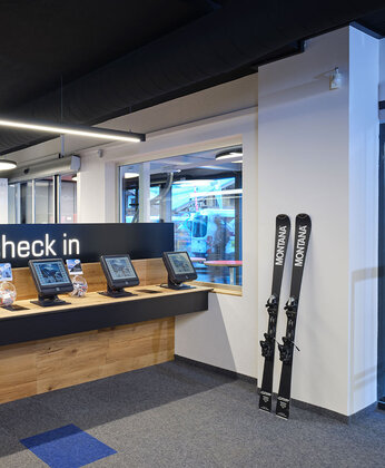 Eine Reihe von Selbstbedienungs-Check-in-Automaten unter einem "Check-in"-Schild in einer modernen, hell erleuchteten Raum. An der Wand steht ein paar Ski.