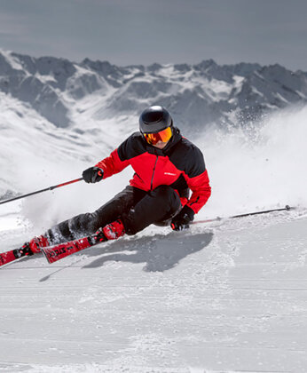 Person beim Skifahren auf einem verschneiten Berghang, mit roter Jacke und schwarzem Helm, mit Bergen im Hintergrund.