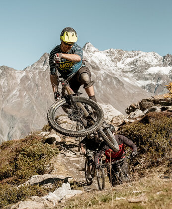 A mountain biker wearing a helmet rides downhill on a forest trail with snow-covered mountains in the background.