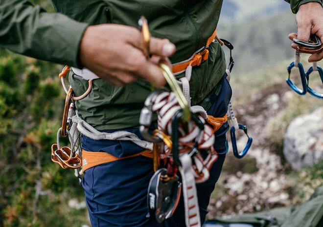 Eine Person in grüner Jacke und Jeans, die eine Kletterausrüstung und Karabiner hält, mit Felsen im Hintergrund.