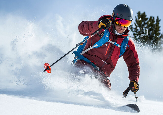 Ein Skifahrer in roter Ausrüstung und mit Helm carvt an einem sonnigen Tag durch den Pulverschnee.