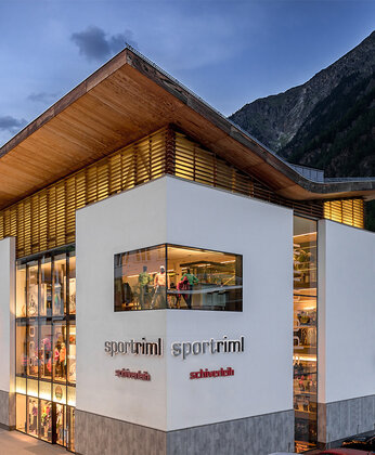 Modernes Sportgeschäft mit großen Glasfenstern, die in der Abenddämmerung beleuchtet werden, vor einer Bergkulisse.