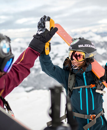 Two skiers in winter gear shake hands on a snowy mountain with skis and snow-covered peaks in the background.