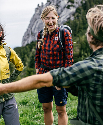 Three people with backpacks are standing and talking outdoors on a meadow with rocky mountains in the background.