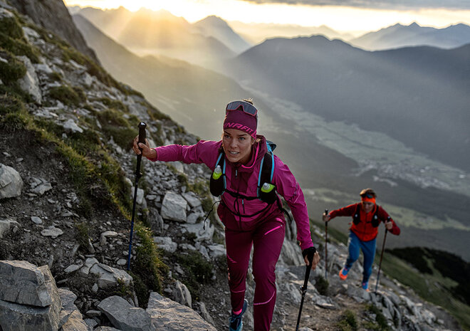 Two hikers climb a rocky mountain trail at sunrise, surrounded by dramatic peaks and a picturesque valley below.