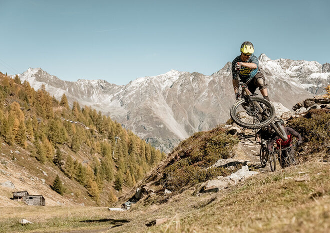 Ein Biker springt über eine unbefestigte Rampe in einer Berglandschaft mit verschneiten Gipfeln und klarem Himmel.
