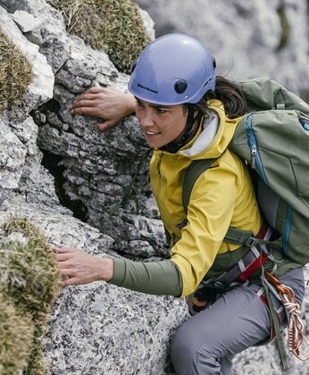 Eine Person, die einen Helm und einen Rucksack trägt, klettert eine felsige Bergwand hinauf und hält sich mit der Hand an den Felsen fest.