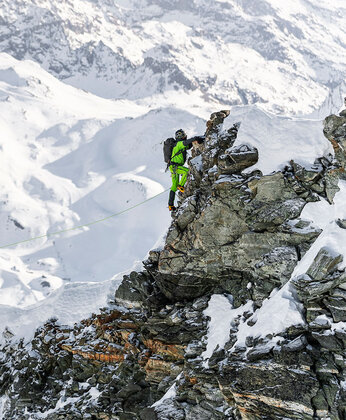 Bergsteiger in leuchtend grüner Ausrüstung beim Aufstieg auf einen zerklüfteten, schneebedeckten Bergkamm mit schneebedeckten Gipfeln im Hintergrund.