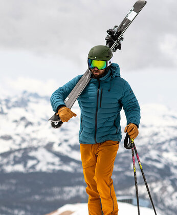 Ein Skifahrer in orangefarbener Hose und blauer Jacke trägt Skier auf der Schulter in einer verschneiten Berglandschaft.