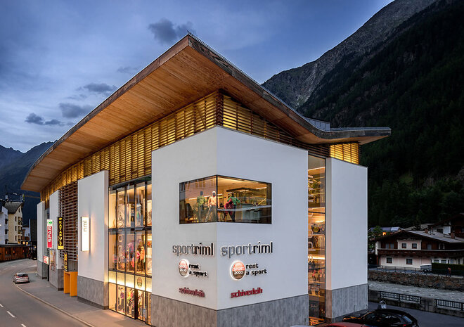 Modernes Sportgeschäft mit großen Glasfenstern, die in der Abenddämmerung beleuchtet werden, vor einer Bergkulisse.