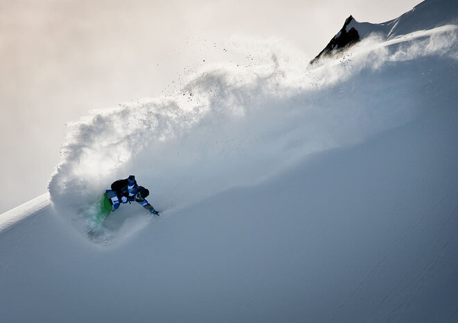 Ein Snowboarder in blauer und grüner Ausrüstung fährt durch den Tiefschnee an einem Berghang und erzeugt eine Schneefontäne.