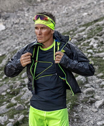 Eine Person, die leuchtend neongelbe Sportkleidung und eine Jacke trägt, steht im Freien auf felsigem Terrain.