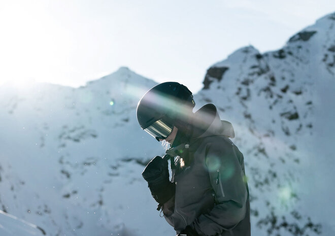 Person in Skikleidung und Helm steht vor verschneiten Bergen im hellen Sonnenlicht.