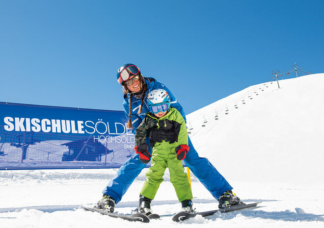 Skilehrer, der einem kleinen Kind auf einer verschneiten Piste unter strahlend blauem Himmel das Skifahren beibringt.