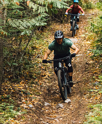 Zwei Personen mit Helmen fahren mit dem Mountainbike einen schmalen Waldweg hinunter, der von Bäumen und Laub umgeben ist.