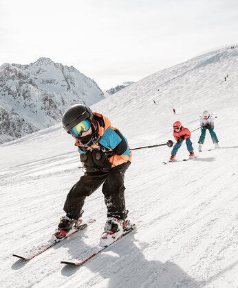 Kind beim Skifahren in den verschneiten Bergen, mit zwei anderen Skifahrern im Hintergrund unter einem hellen Himmel.