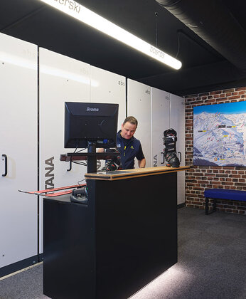 Eine Person steht hinter einem modernen schwarzen Empfangstresen mit einem Computer in einem eleganten, gut beleuchteten Raum mit Skischuh am Tresen.