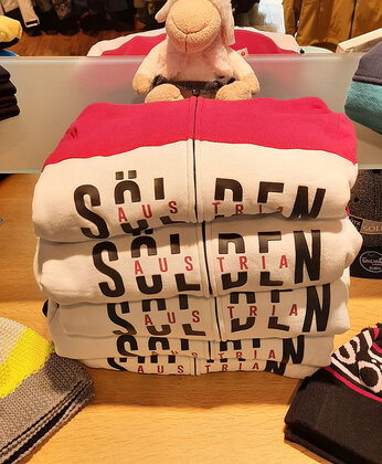 Vier gefaltete Sweatshirts mit der Aufschrift Sölden auf einem Tisch, auf dem ein lächelndes Plüschschaf sitzt.